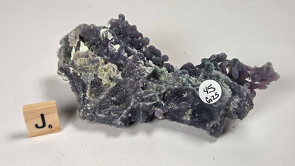 A purple and gray mineral specimen with a rough, crystalline texture, displayed on a light gray background next to a small wooden tile with a 'J' and a white inventory sticker.