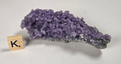 A purple crystal cluster mineral specimen with a branching base, displayed on a light gray surface next to a wooden Scrabble tile showing the letter K.