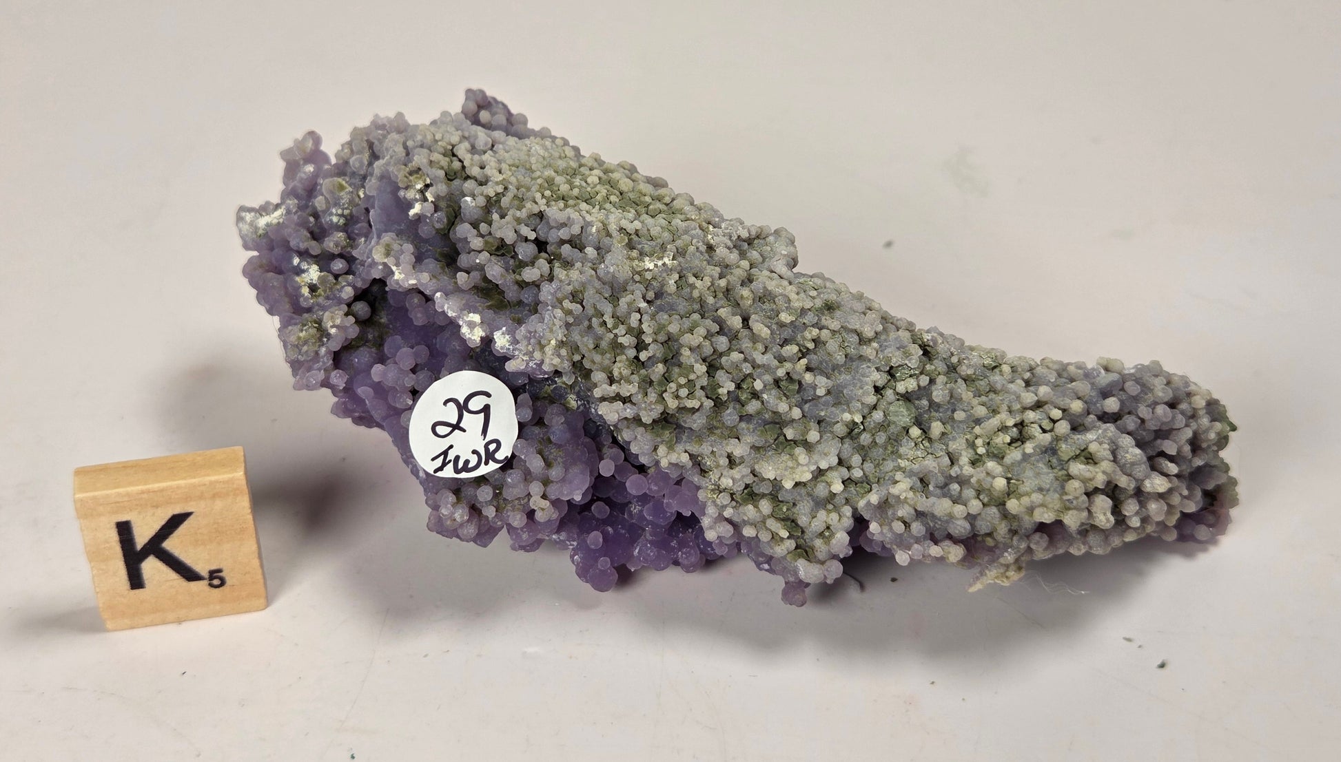 A natural amethyst crystal cluster with a lavender-purple to pale gray-green color transition, displayed on a light off-white background next to a small wooden letter tile and a white inventory sticker.