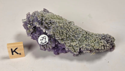 A natural amethyst crystal cluster with a lavender-purple to pale gray-green color transition, displayed on a light off-white background next to a small wooden letter tile and a white inventory sticker.