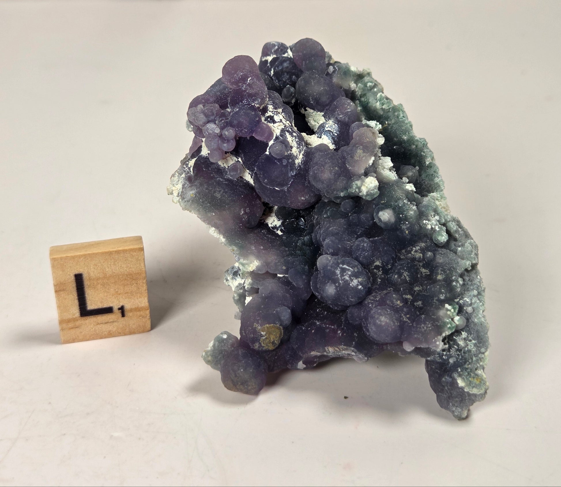 A purple and blue-green amethyst crystal cluster with white calcite, shown next to a small wooden letter 'L' block on a light gray surface.