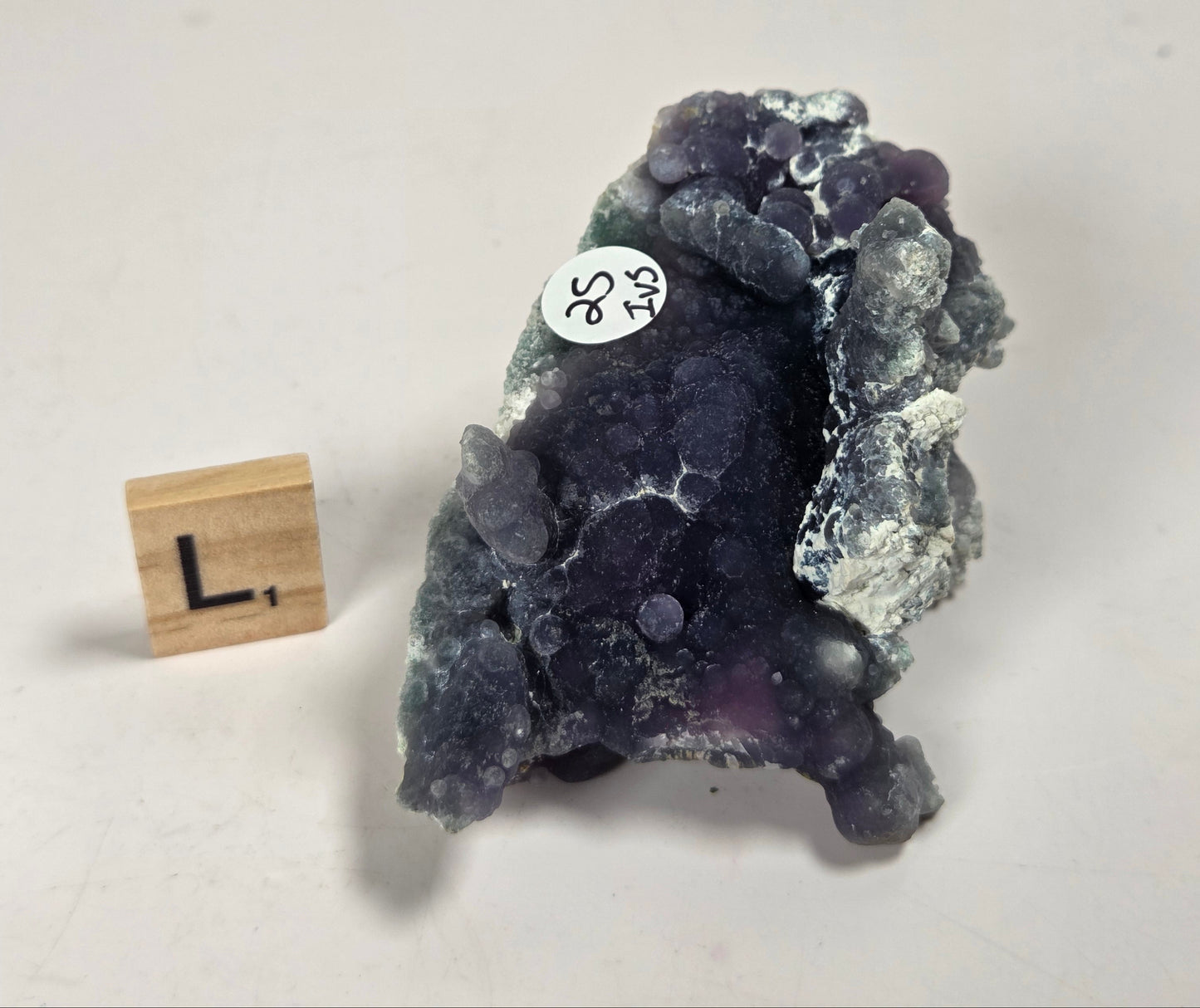 A raw amethyst geode specimen with dark purple and gray crystalline formations, shown next to a small wooden letter block displaying 'L.' on a plain off-white background.
