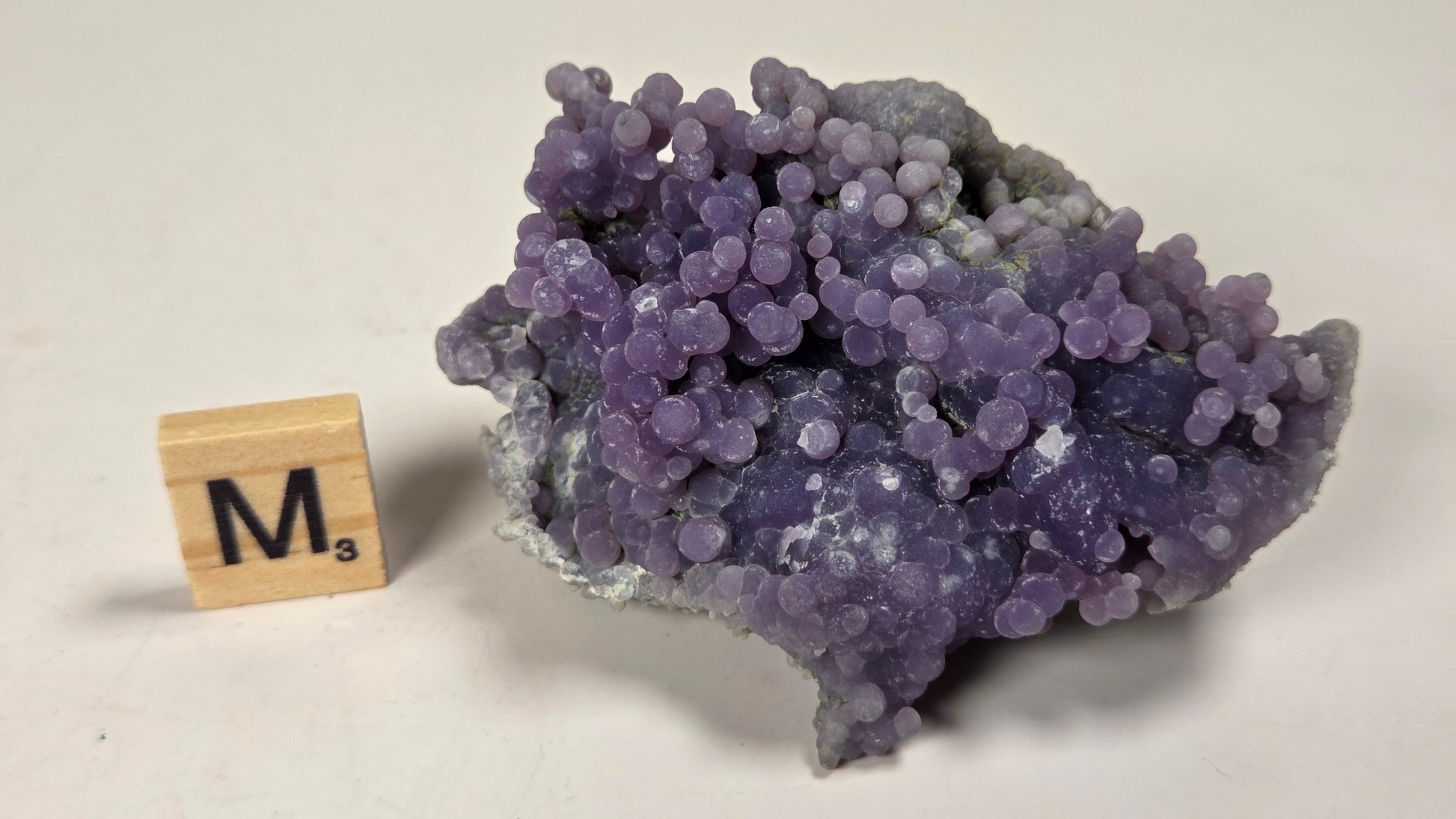 A purple amethyst crystal cluster with rounded, bubble-like formations on a light gray background, next to a wooden Scrabble tile showing the letter 'M'.