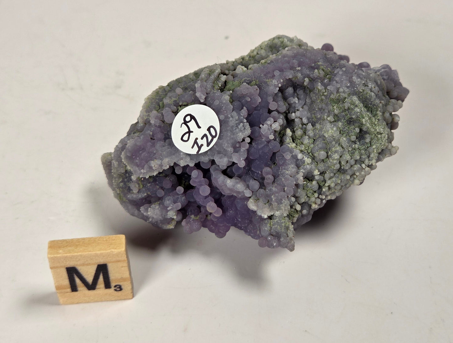 A purple and green crystalline mineral specimen with a small white label reading 'J9 210', photographed on a light neutral surface next to a small wooden block marked 'M'.