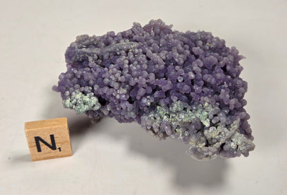 A purple and lavender crystalline mineral specimen with light greenish-gray areas, displayed on a plain off-white background next to a small wooden letter block showing the letter 'N'.