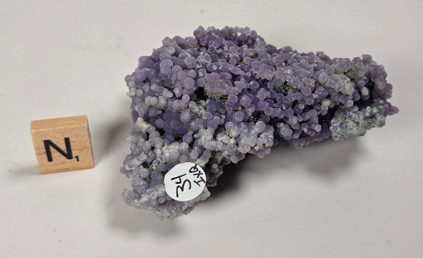 A purple mineral specimen with a textured, bubble-like crystalline structure, shown next to a wooden letter block and a handwritten label on a light gray background.