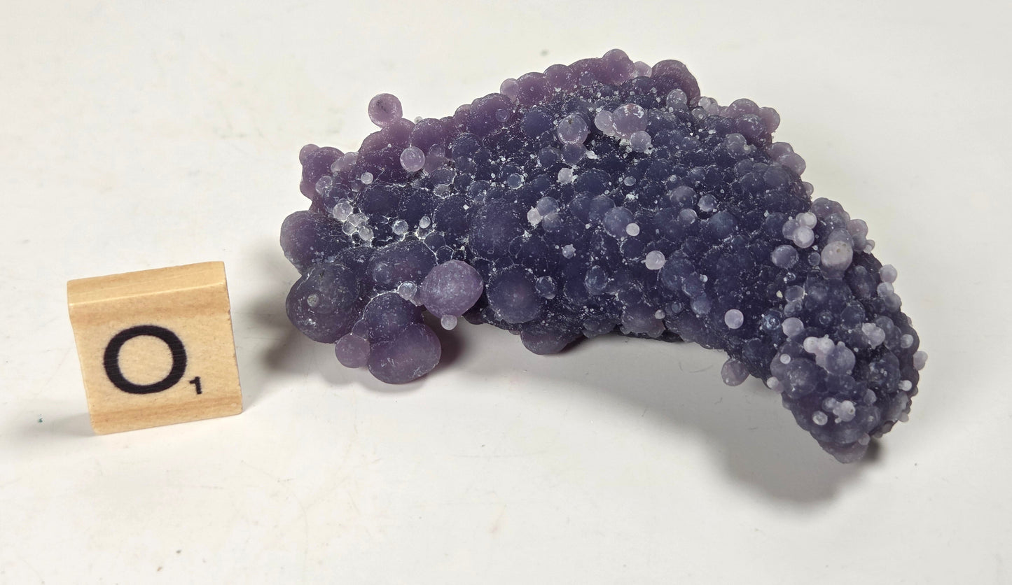 A purple and violet amethyst crystal cluster with rounded, bubble-like formations, shown next to a small wooden Scrabble tile with the letter 'O' on a plain off-white background.