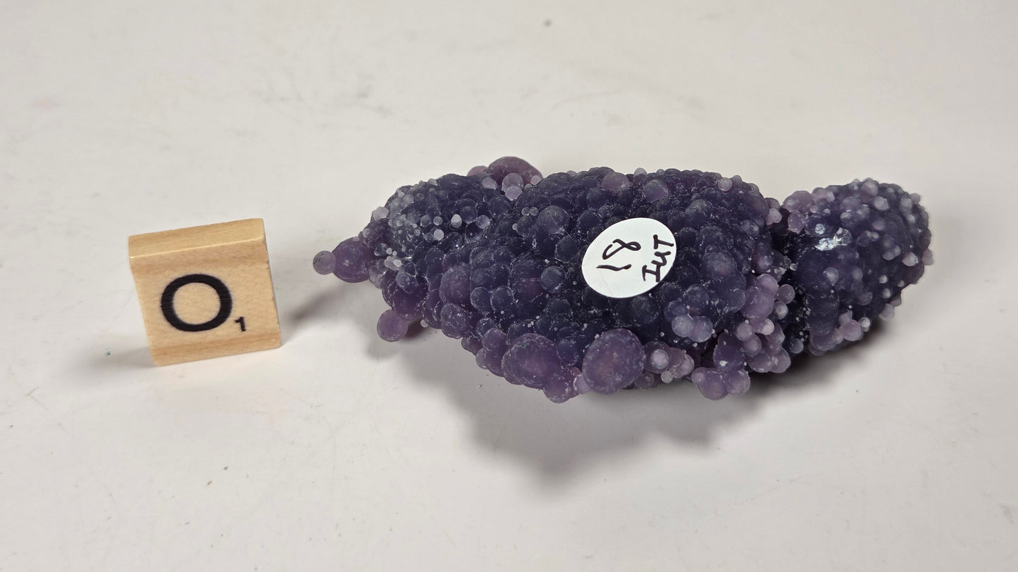 A purple amethyst geode specimen with a bubbly, bumpy crystal texture, displayed on a light gray surface next to a wooden Scrabble tile showing the letter 'O' and a small white label.