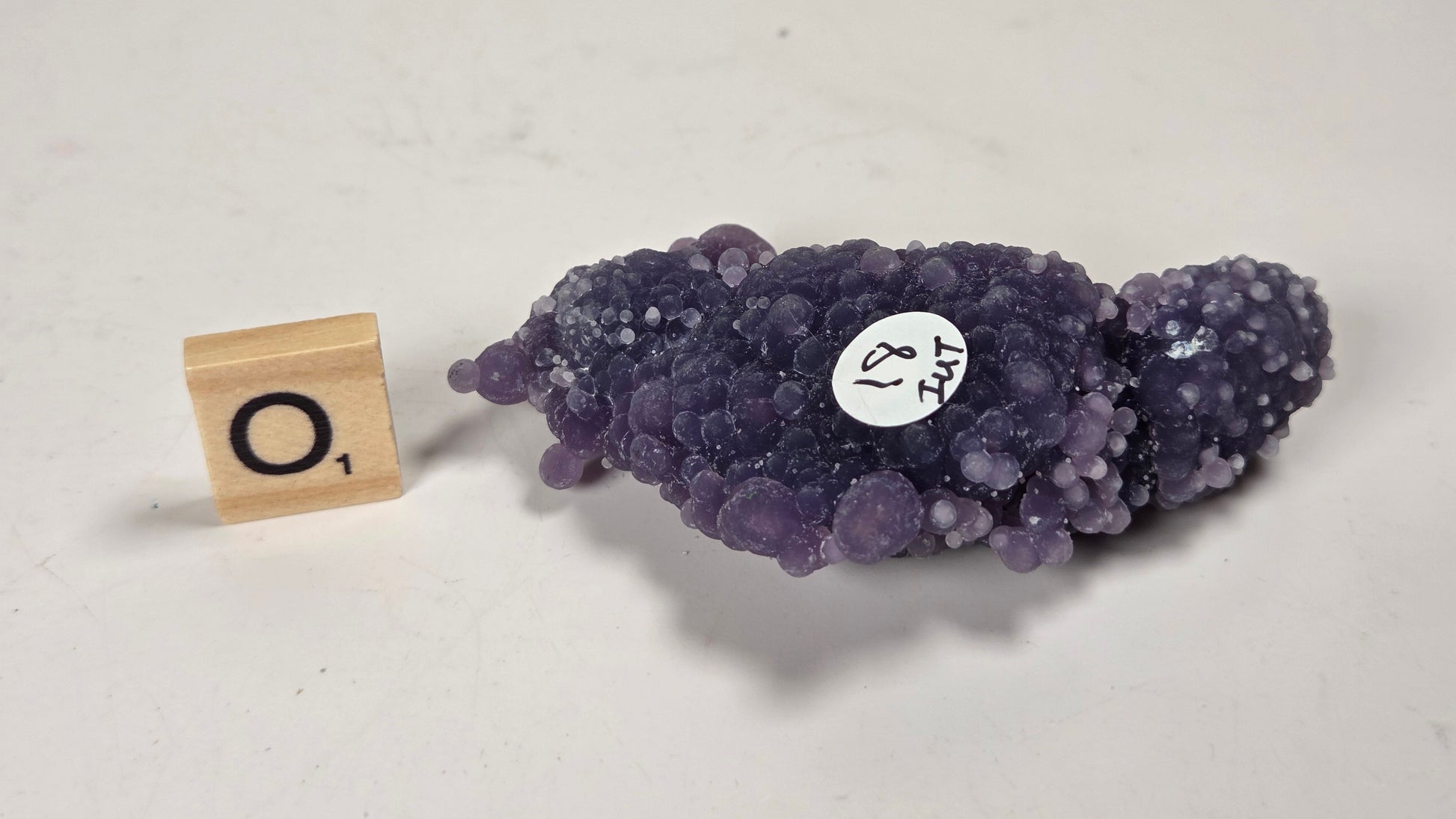 A purple amethyst geode specimen with a bubbly, bumpy crystal texture, displayed on a light gray surface next to a wooden Scrabble tile showing the letter 'O' and a small white label.
