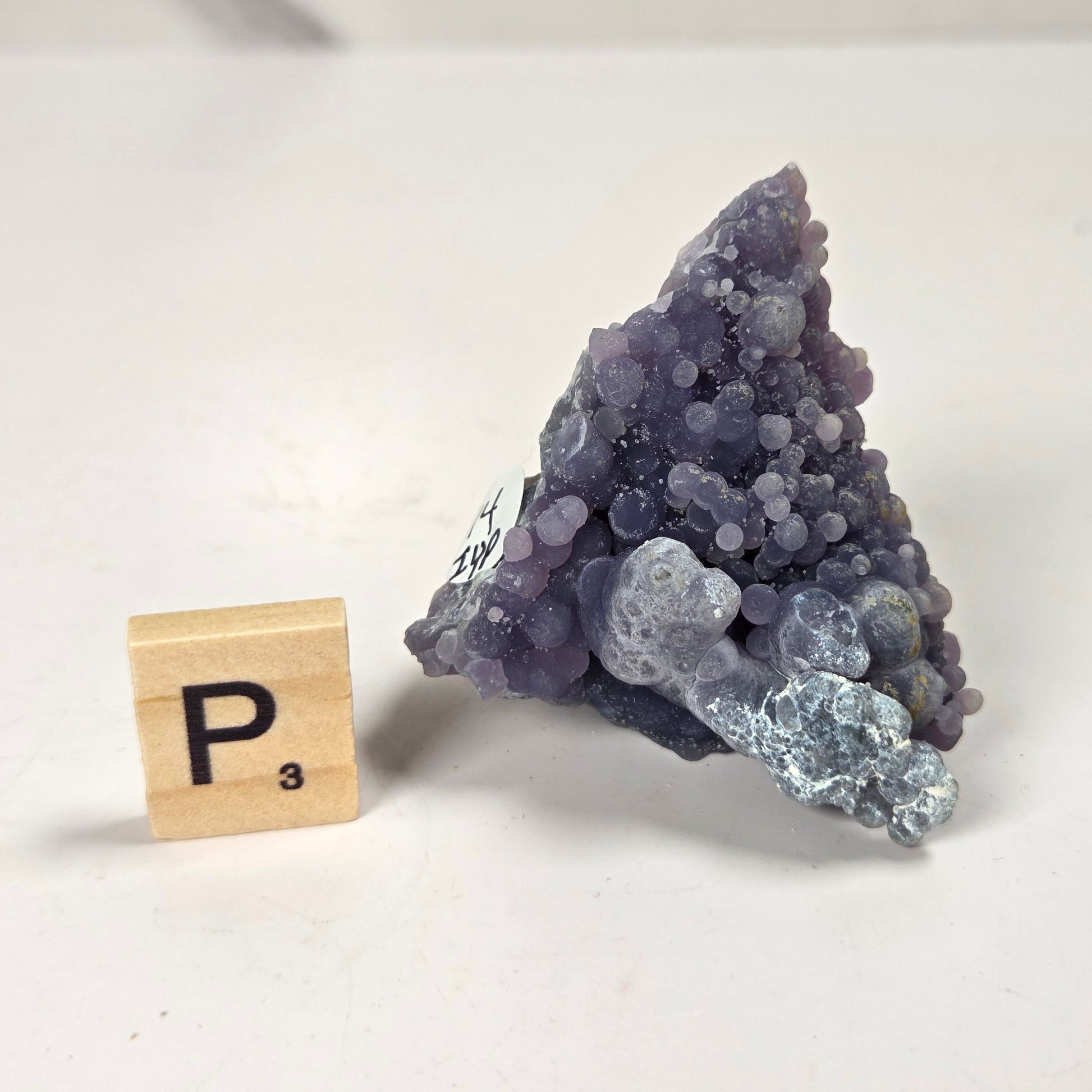 A triangular cluster of purple and lavender amethyst geode crystals with a light blue agate section, displayed next to a wooden Scrabble tile spelling 'P3' on a plain white surface.