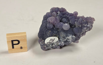 A purple mineral specimen with clustered rounded crystal formations, shown next to a wooden Scrabble tile and a small white label, on a plain off-white background.