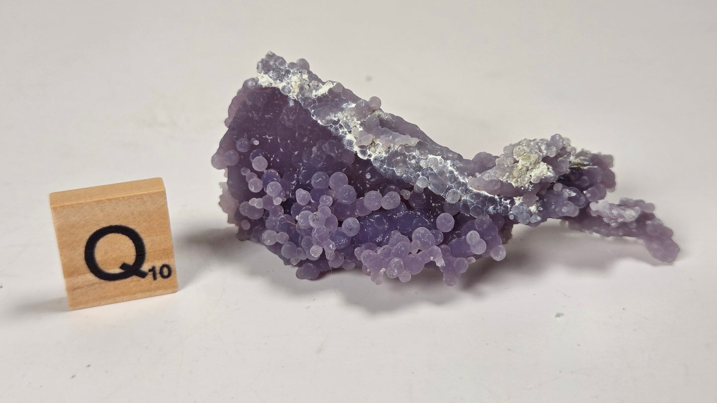 A purple amethyst geode specimen with silver-white metallic crystal formations, displayed next to a wooden Scrabble tile showing 'Q-10' on a plain off-white background.