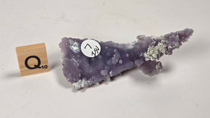 A purple amethyst geode specimen with white calcite crystals, shown next to a small wooden block scale marker on a plain off-white background.