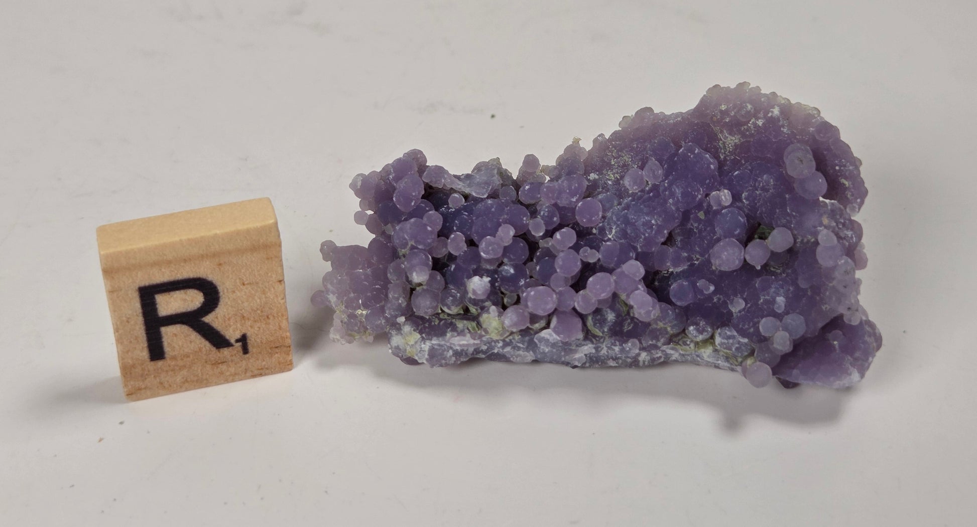 A small cluster of purple crystalline mineral or gemstone specimen shown next to a wooden letter tile with the letter R on a plain light background.