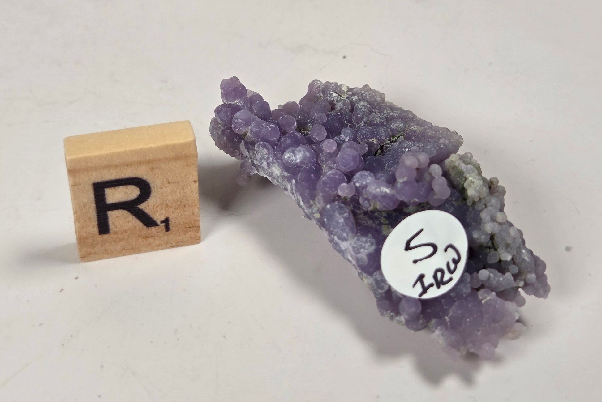 A small purple mineral or crystal specimen with a rough, clustered texture, shown next to a wooden letter block with the letter 'R' and a white circular label reading 'S 123', on a plain off-white surface.