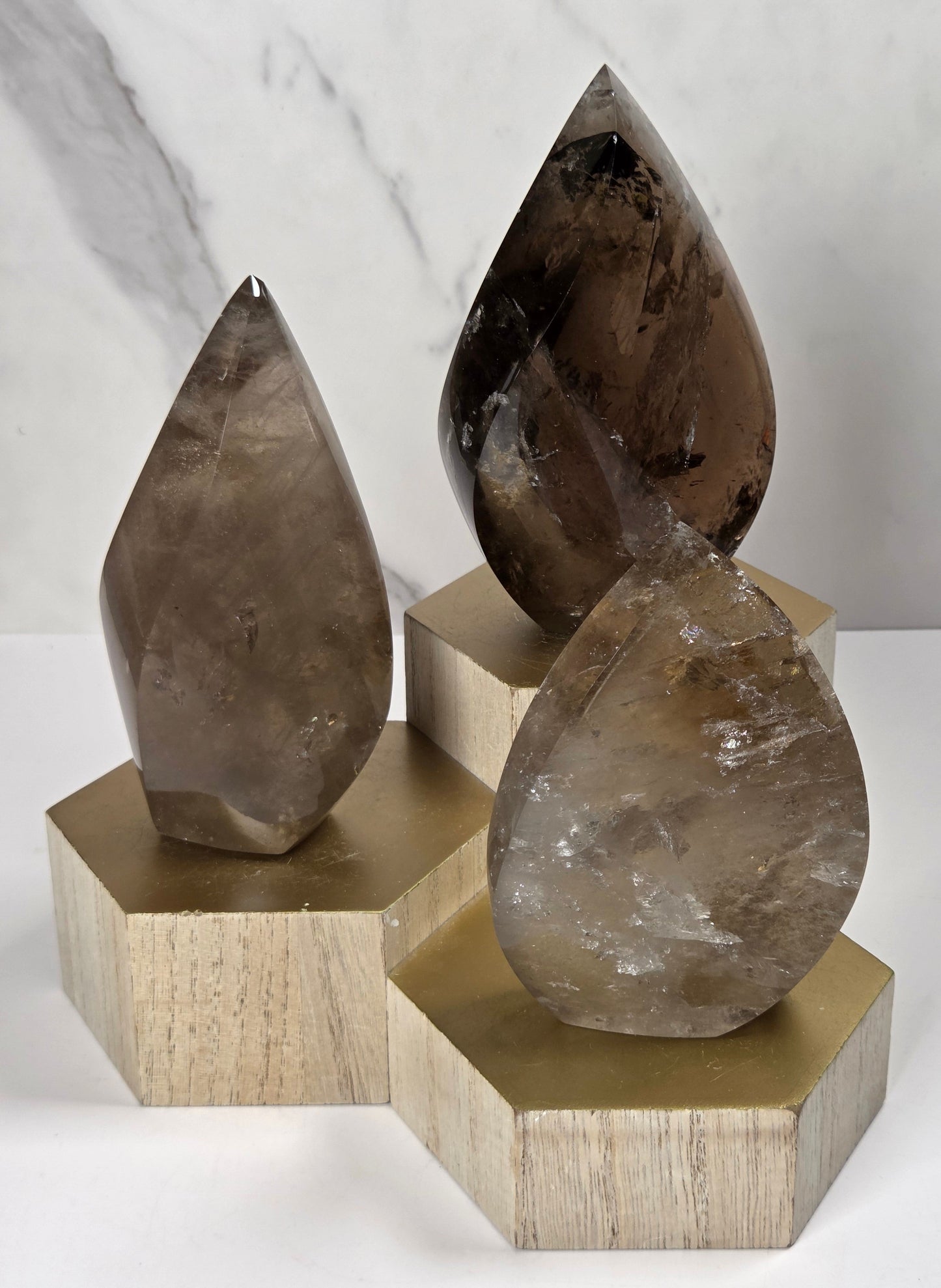 Three polished smoky quartz crystal points of varying sizes, each resting on a hexagonal light wood base, arranged against a white marble background.