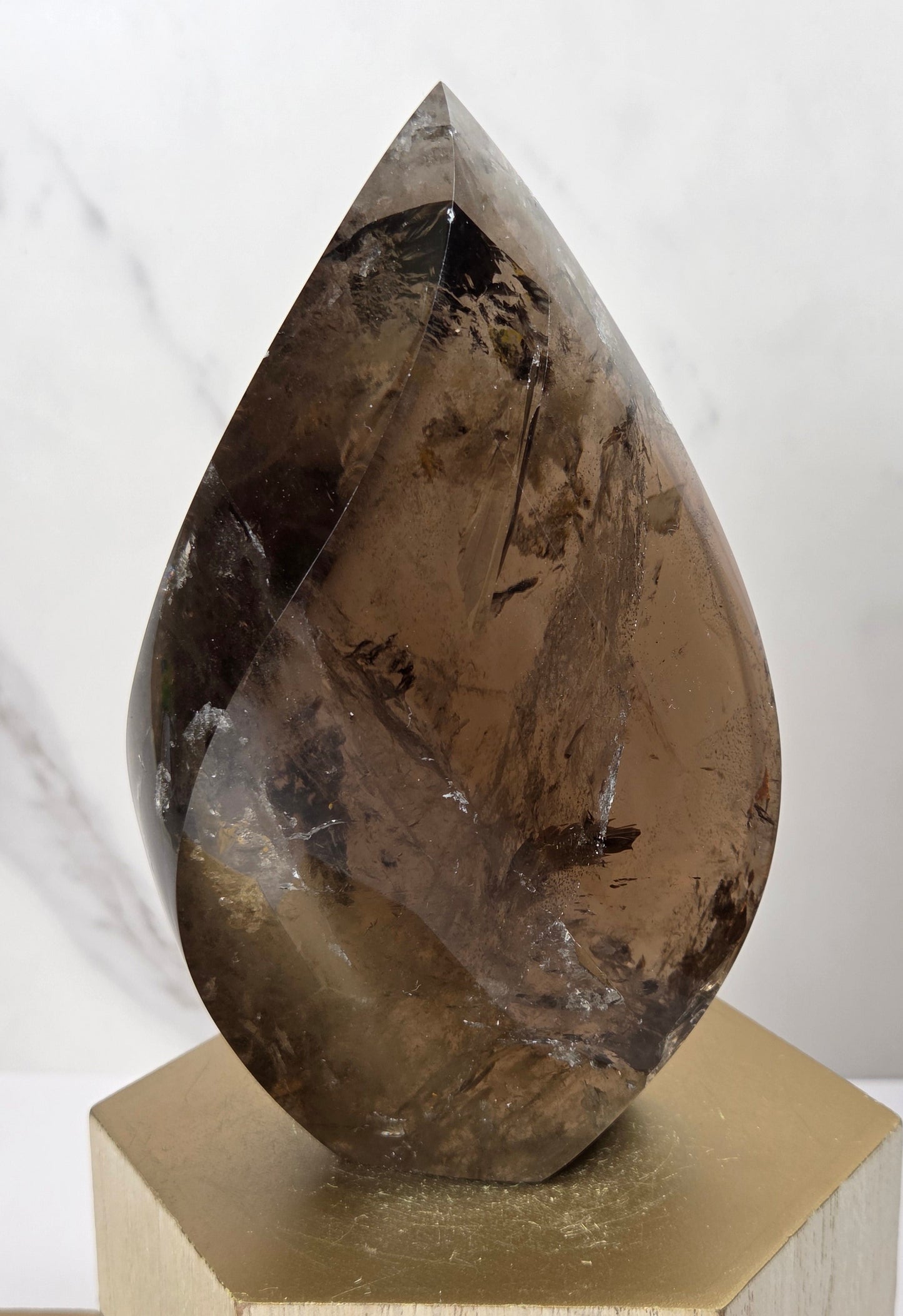 A teardrop-shaped smoky quartz crystal point with natural inclusions, displayed on a gold hexagonal pedestal against a light marble background.