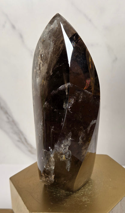 A faceted smoky quartz crystal point resting on a hexagonal gold metal and light wood display stand, set against a white marble-patterned background.