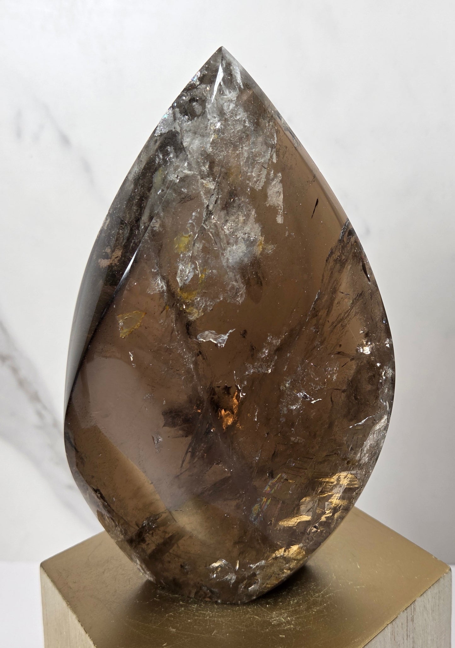 A polished teardrop-shaped smoky quartz crystal point resting on a gold hexagonal display block against a light gray marble background.