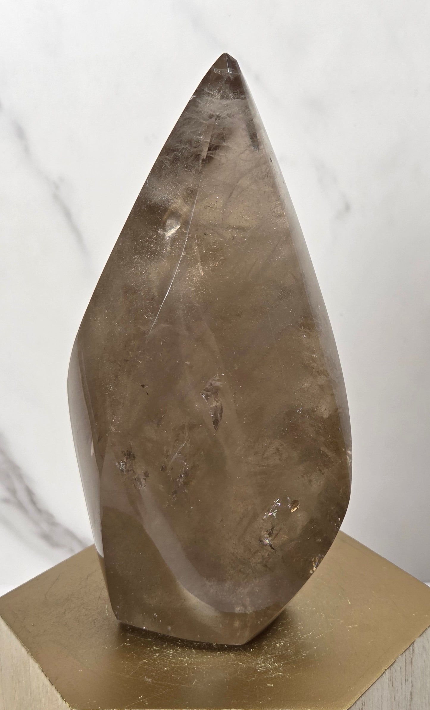 A polished natural crystal point, likely smoky quartz, standing upright on a gold and light wood hexagonal pedestal against a white marble-patterned background.