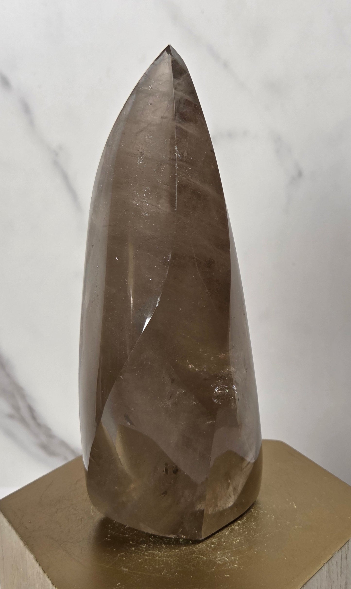 A faceted smoky quartz crystal point displayed on a gold hexagonal metal stand against a white marble-patterned background.