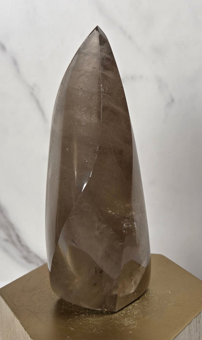 A faceted smoky quartz crystal point displayed on a gold hexagonal metal stand against a white marble-patterned background.