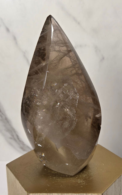 A teardrop-shaped smoky quartz crystal point resting on a gold hexagonal metal pedestal against a light gray marble background.