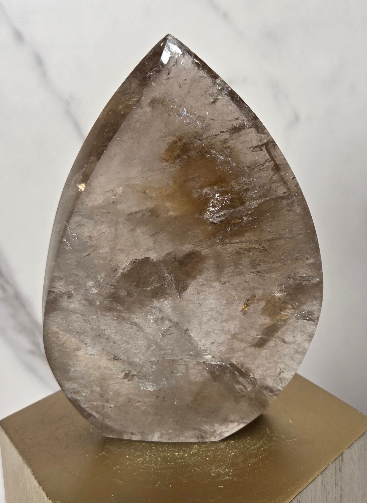 A teardrop-shaped polished smoky quartz crystal sculpture displayed on a gold hexagonal pedestal against a light marble background.