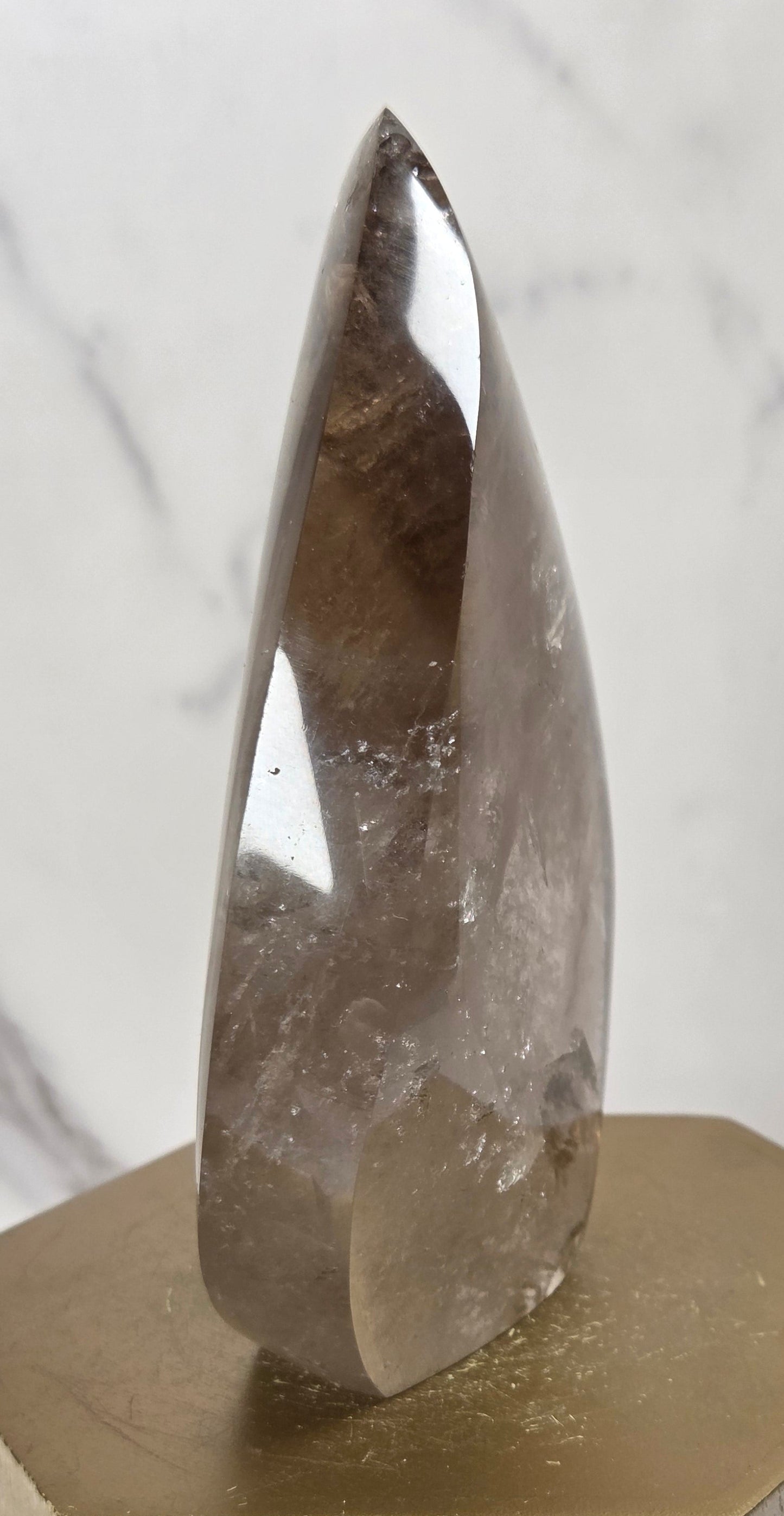 A polished smoky quartz crystal point displayed on a hexagonal wooden block with a gold-finished top surface, set against a light gray marble-patterned background.