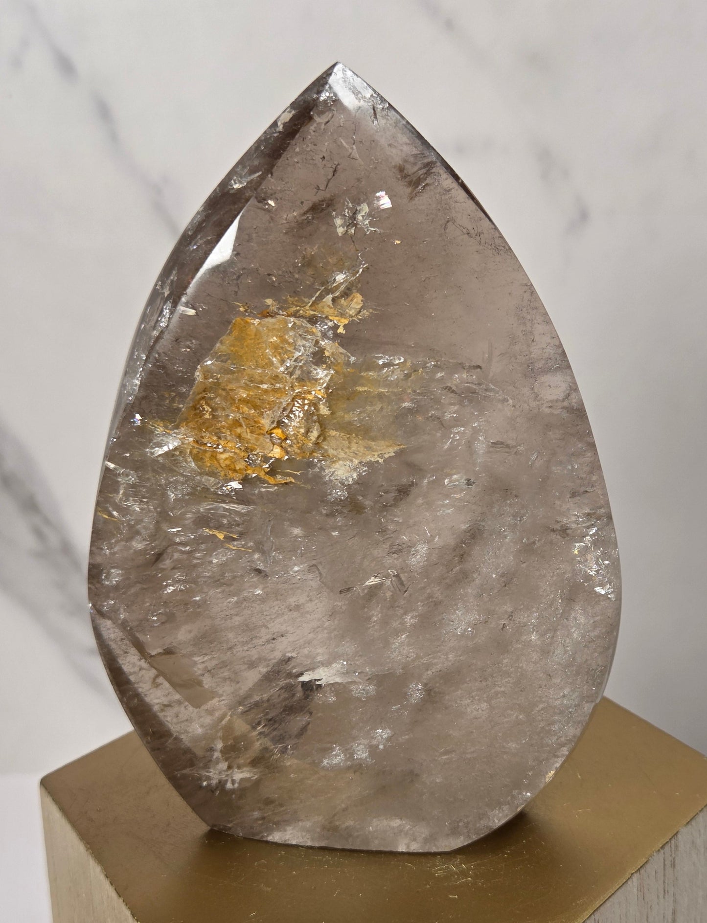 A teardrop-shaped clear quartz crystal with natural inclusions and a gold leaf inlay, displayed on a gold hexagonal pedestal against a light gray marble background.