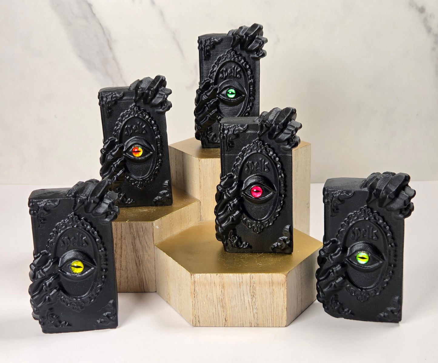 Five black rectangular decorative objects with ornate sculpted sides and a circular inset featuring a single colored eye, displayed on wooden blocks against a light marble-like background.