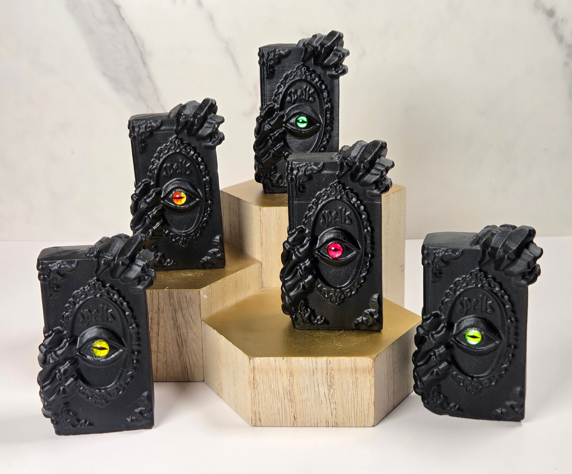 Five black rectangular decorative objects with ornate sculpted sides and a circular inset featuring a single colored eye, displayed on wooden blocks against a light marble-like background.