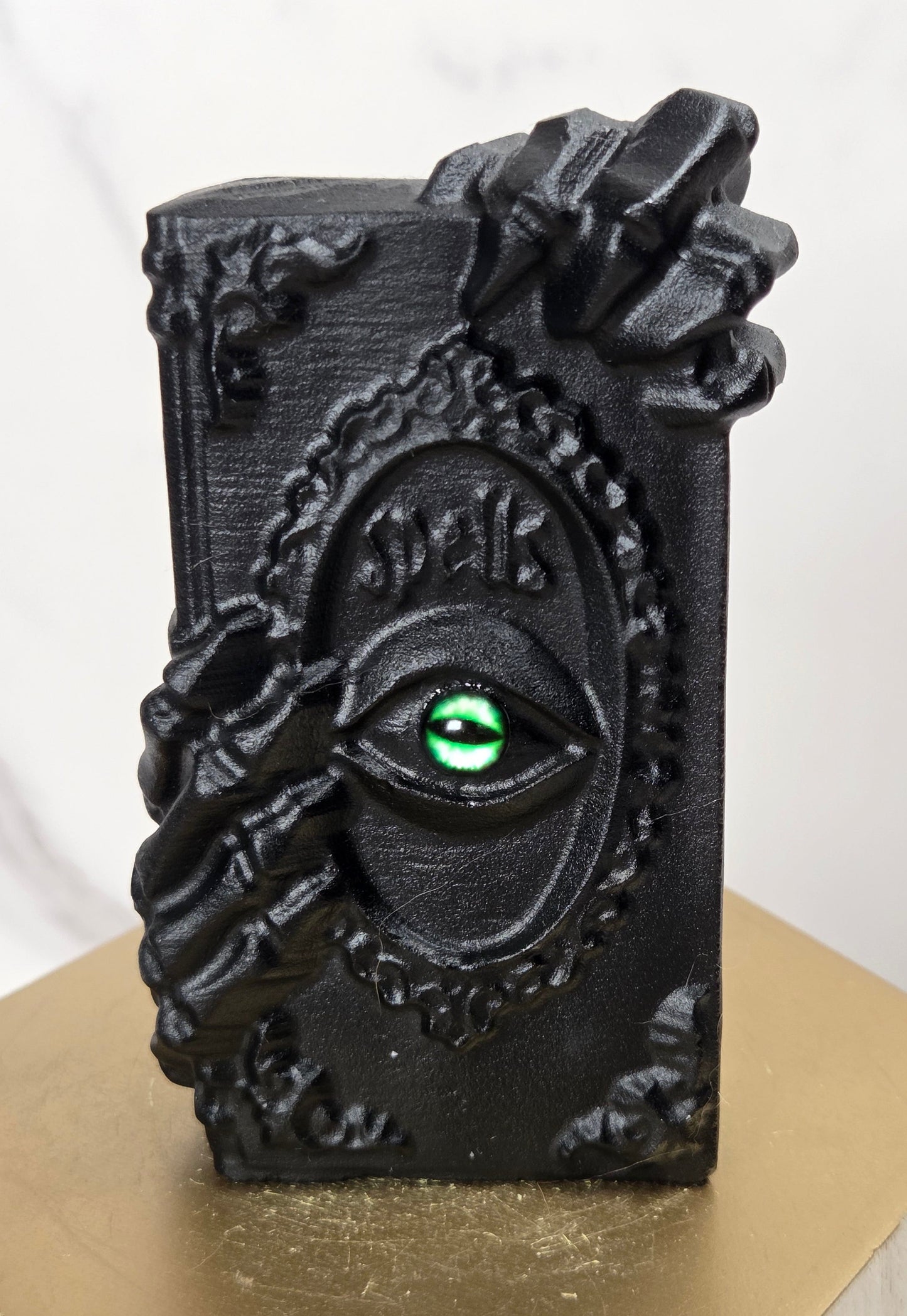 A black gothic-style decorative plaque or box featuring a raised eye motif with a green iris and the word 'Eyes' in script, displayed on a gold-colored hexagonal pedestal against a light marble background.