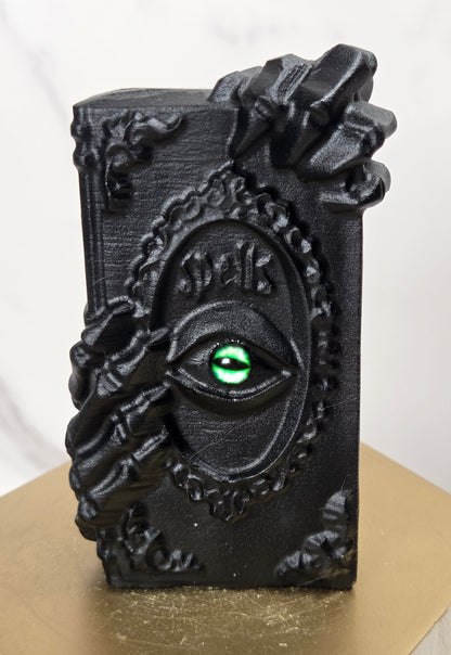 A black gothic-style decorative plaque or box featuring a raised eye motif with a green iris and the word 'Eyes' in script, displayed on a gold-colored hexagonal pedestal against a light marble background.