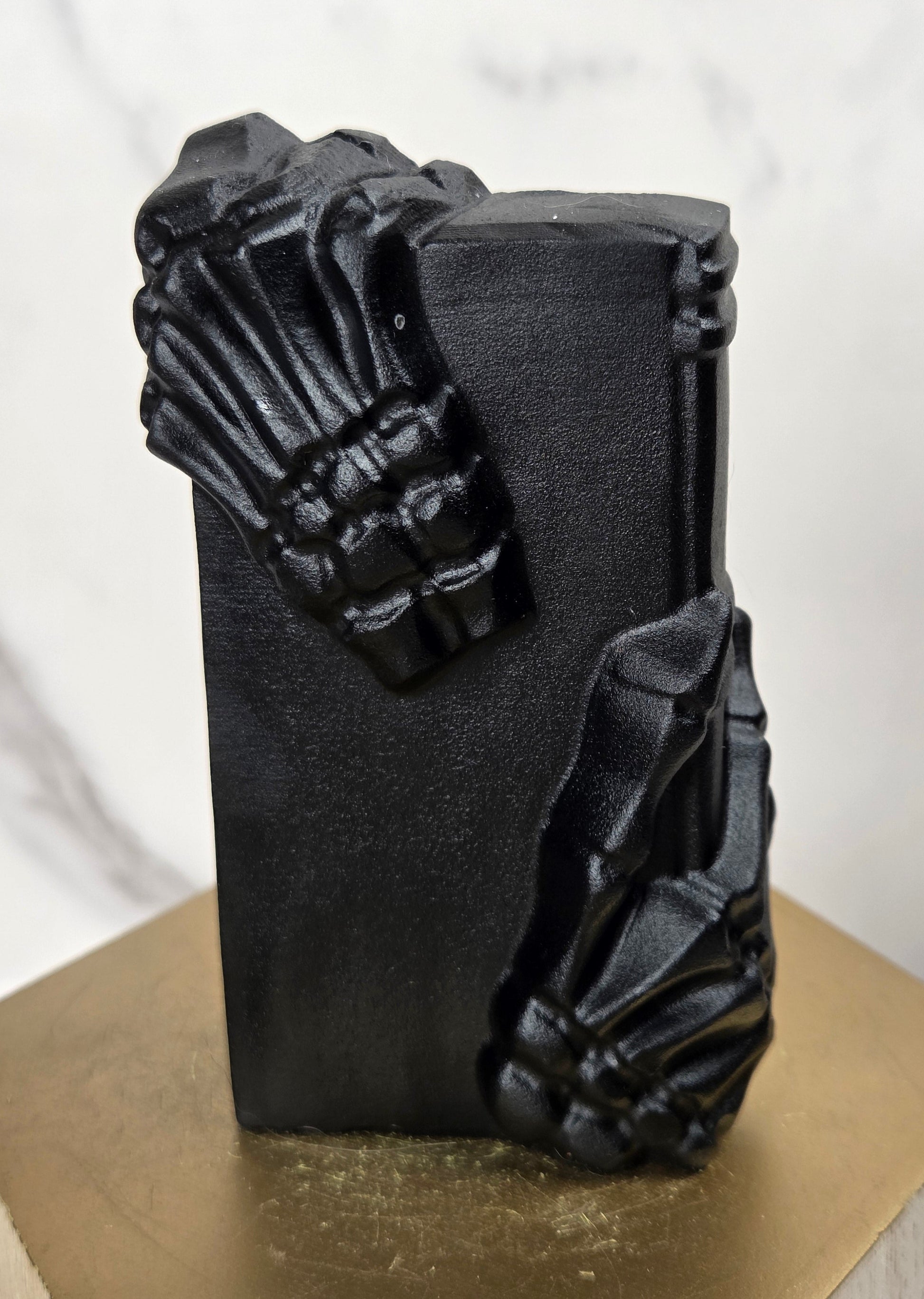 A black rectangular decorative object with sculpted skeletal hands gripping the sides, displayed on a gold hexagonal pedestal against a light marble-patterned background.
