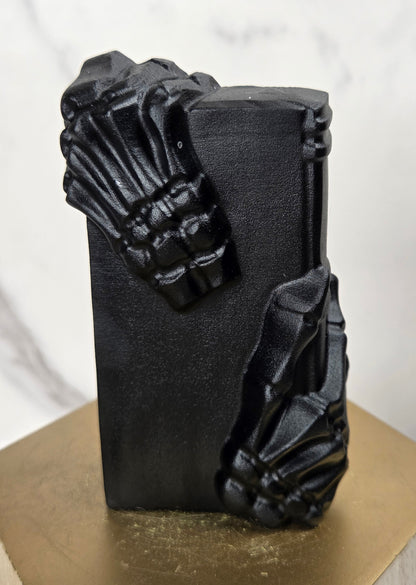 A black rectangular decorative object with sculpted skeletal hands gripping the sides, displayed on a gold hexagonal pedestal against a light marble-patterned background.