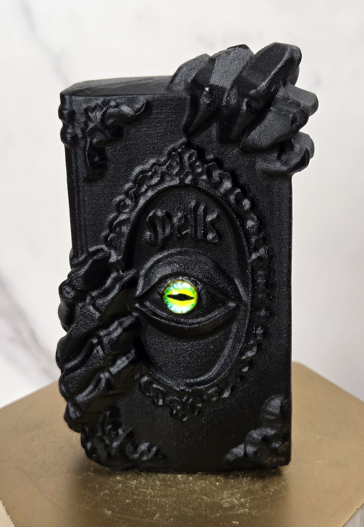 A black gothic decorative object featuring a raised eye with a glowing green-yellow iris and ornate claw-like hand details, displayed on a gold hexagonal pedestal against a light marble background.