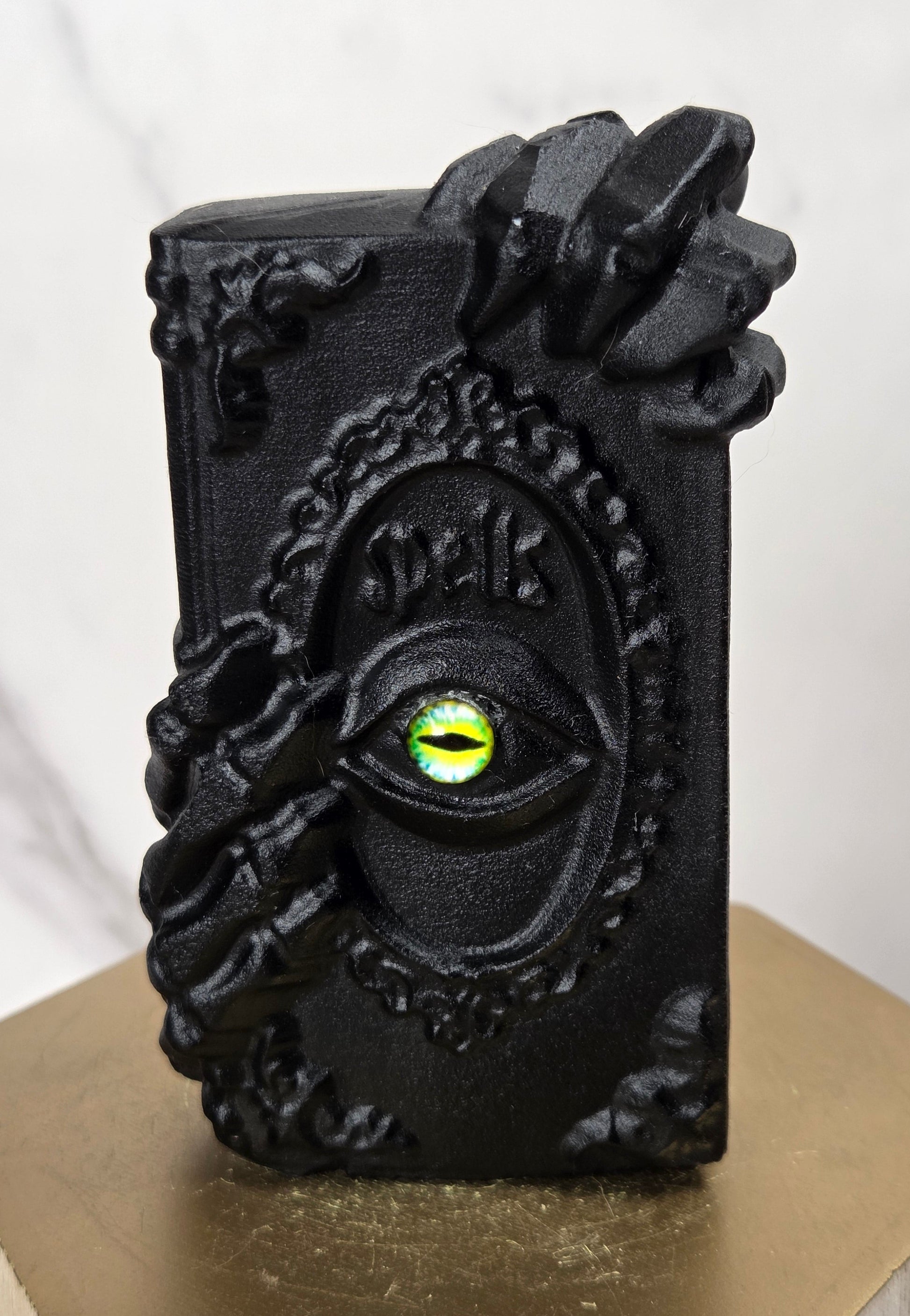 A black gothic decorative object featuring a raised eye with a glowing green-yellow iris and ornate claw-like hand details, displayed on a gold hexagonal pedestal against a light marble background.