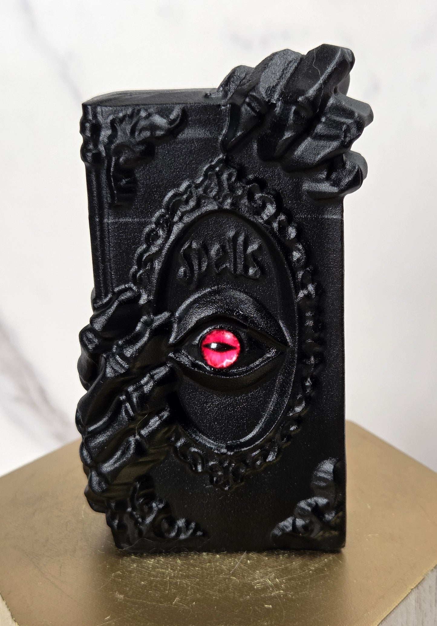 A black decorative book-shaped object with ornate hand carvings and a red eye motif on the front, displayed on a gold hexagonal pedestal against a light marble background.