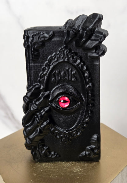 A black decorative book-shaped object with ornate hand carvings and a red eye motif on the front, displayed on a gold hexagonal pedestal against a light marble background.