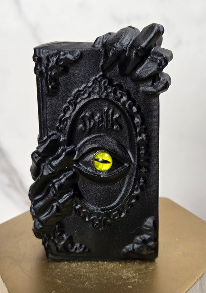 A black decorative book-shaped object with skeletal hands gripping the sides and a yellow eye motif on the front, displayed on a gold pedestal against a light marble background.