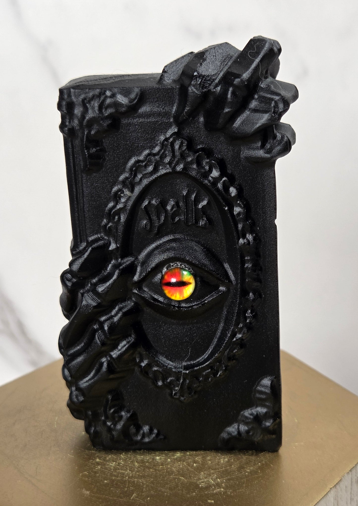 A black gothic-style decorative object shaped like a book or box with skeletal hands and an eye motif, featuring a red and yellow gem-like center, displayed on a gold hexagonal pedestal against a light marble background.