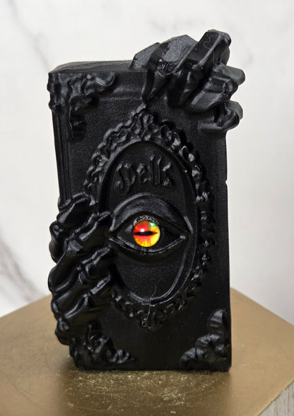 A black gothic-style decorative object shaped like a book or box with skeletal hands and an eye motif, featuring a red and yellow gem-like center, displayed on a gold hexagonal pedestal against a light marble background.