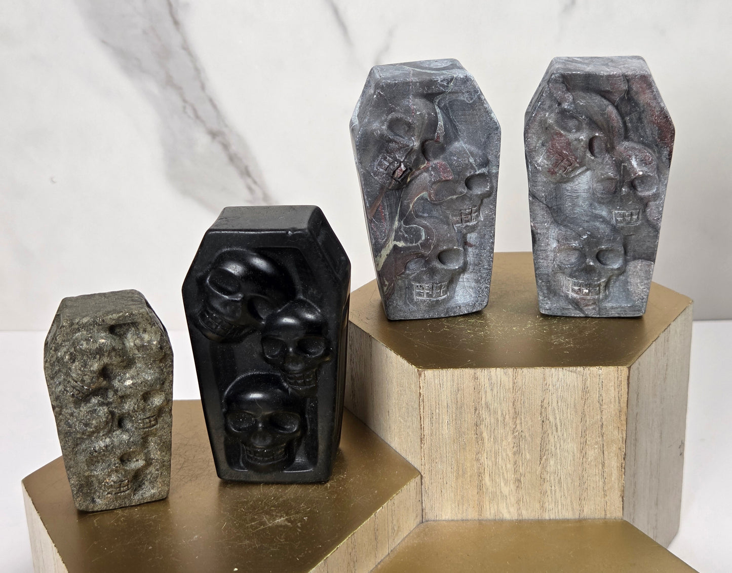 Four carved stone coffin-shaped sculptures with skull details, displayed on geometric gold and wood pedestals against a light marble background.