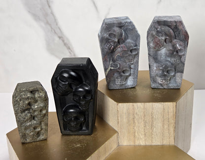 Four carved stone coffin-shaped sculptures with skull details, displayed on geometric gold and wood pedestals against a light marble background.