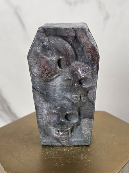 A small carved stone figurine shaped like a coffin with three stacked skulls, displayed on a gold hexagonal pedestal against a light marble background.