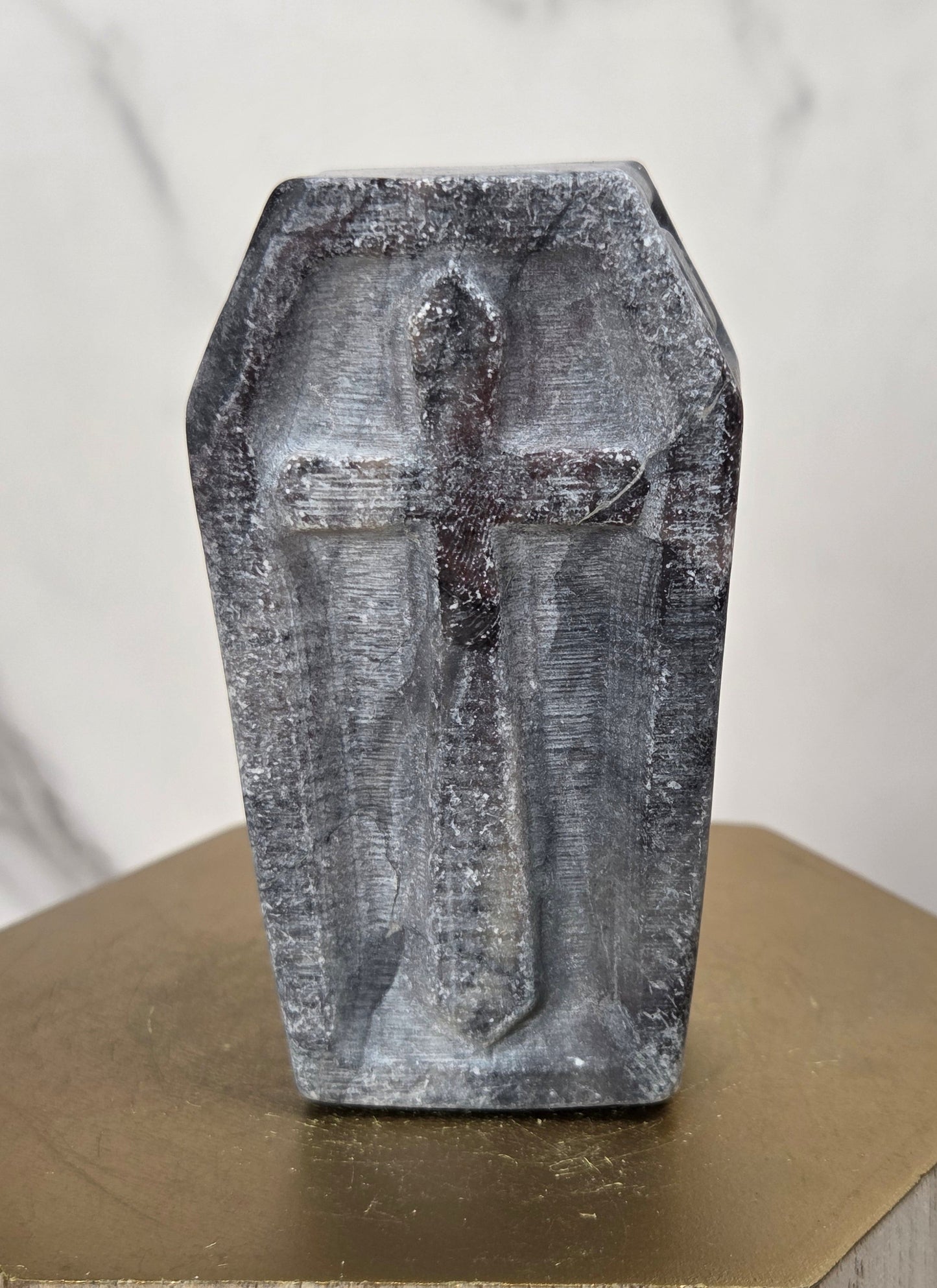A small gray stone or concrete coffin-shaped cross sculpture standing upright on a gold-toned hexagonal pedestal against a light marble-patterned background.