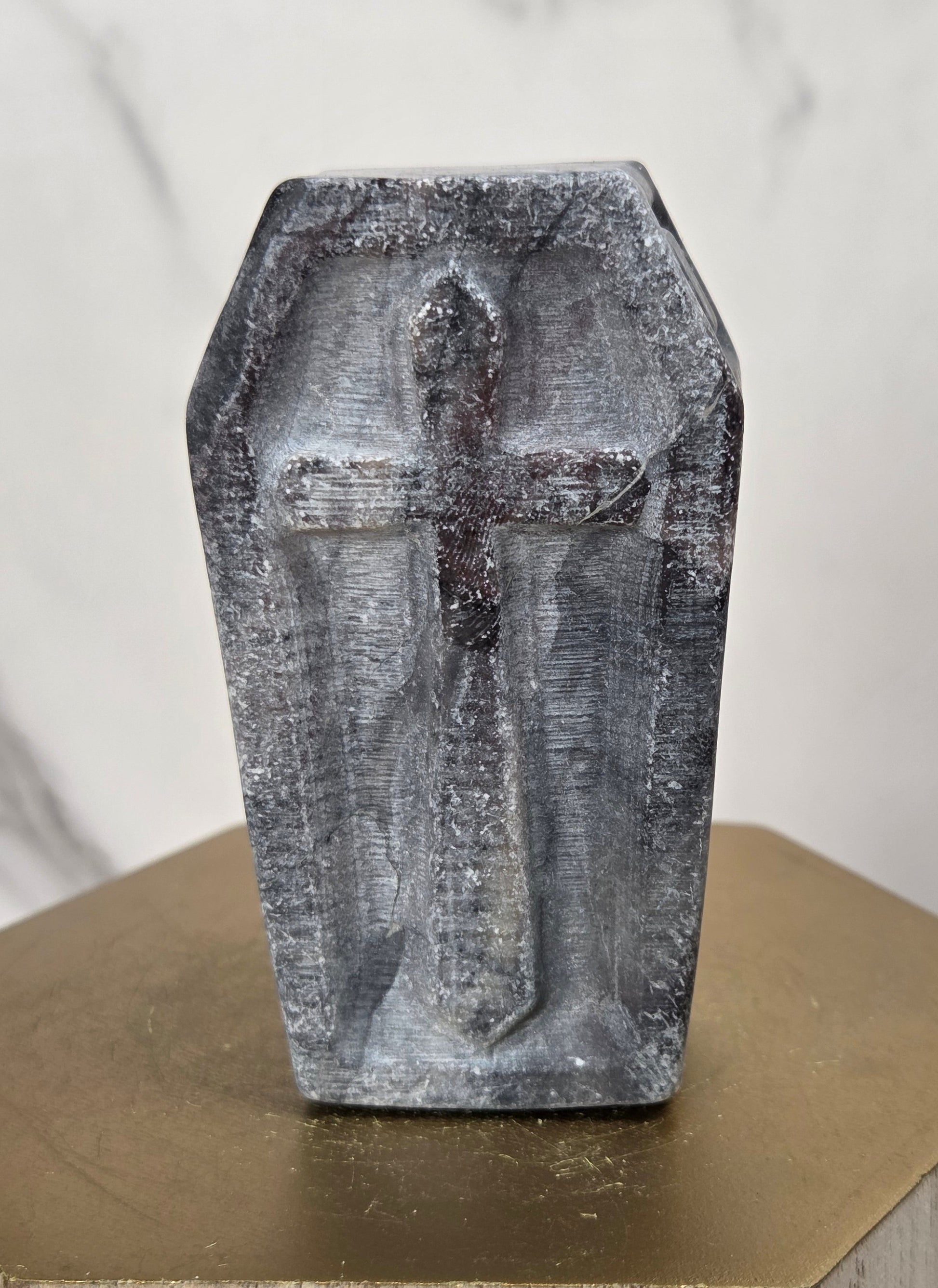 A small gray stone or concrete coffin-shaped cross sculpture standing upright on a gold-toned hexagonal pedestal against a light marble-patterned background.