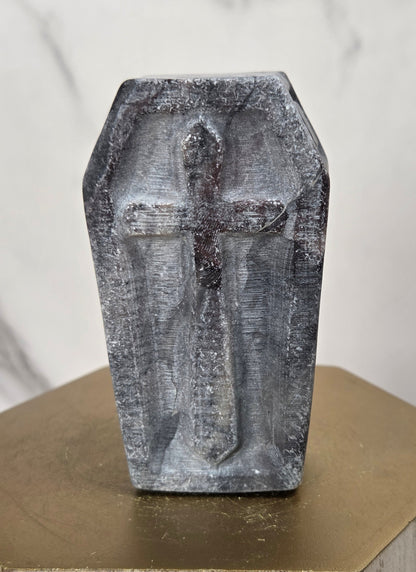A small gray stone or concrete coffin-shaped cross sculpture standing upright on a gold-toned hexagonal pedestal against a light marble-patterned background.
