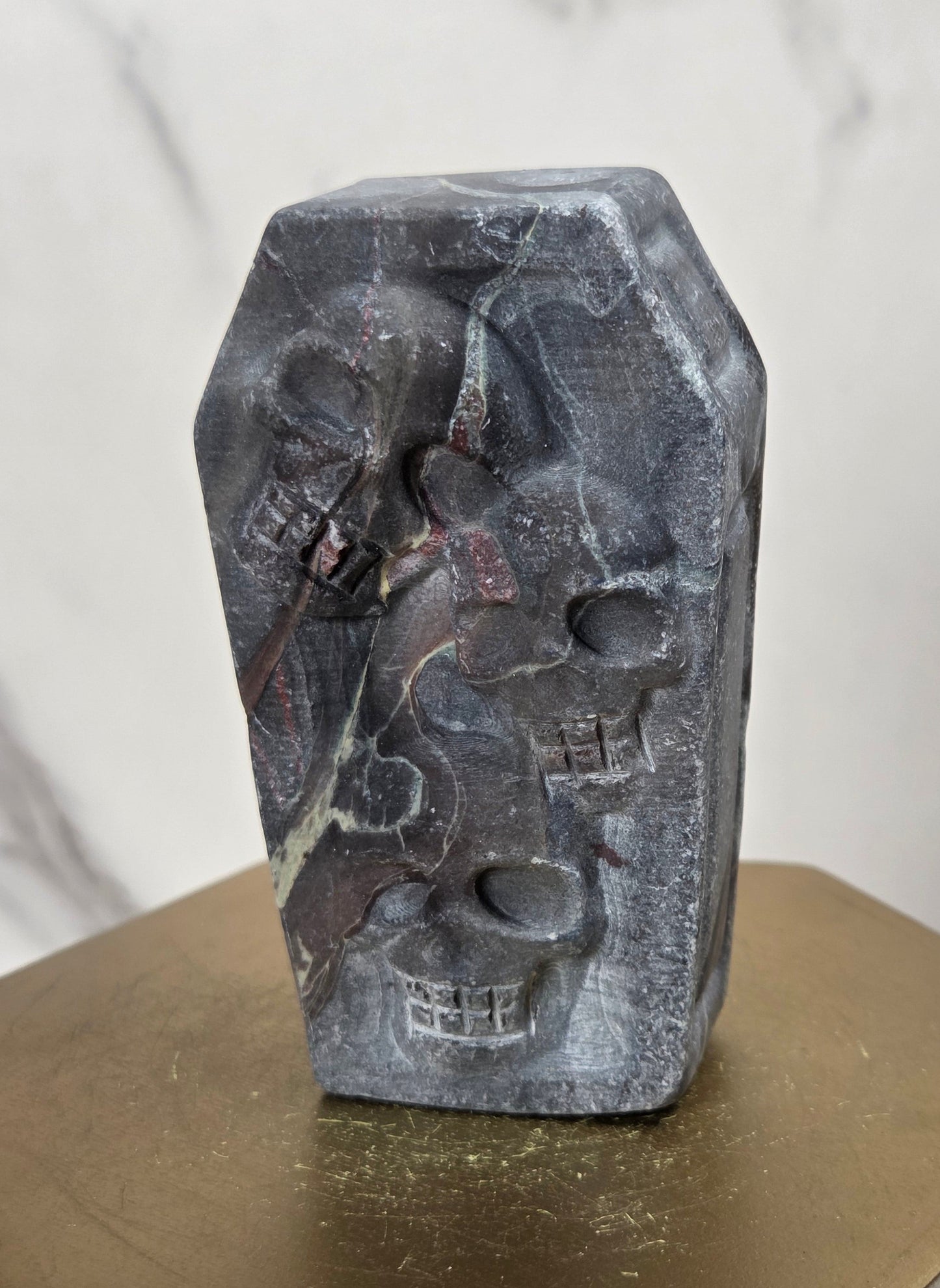 A small carved stone object shaped like a coffin, featuring raised skull details, displayed on a gold-toned hexagonal pedestal against a light marble-patterned background.